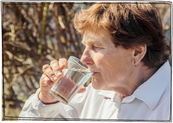 Eine Frau um die 60 vor einem Baum: Sie trinkt ein Glas Wasser.