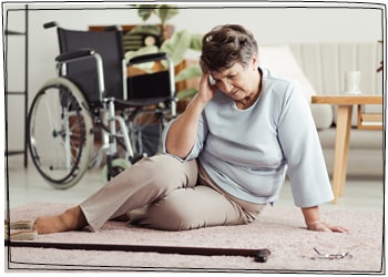 Eine ältere Dame ist gestürzt und sitzt benommen auf einem Wohnzimmerteppich. Hinter ihr steht ein Rollstuhl.